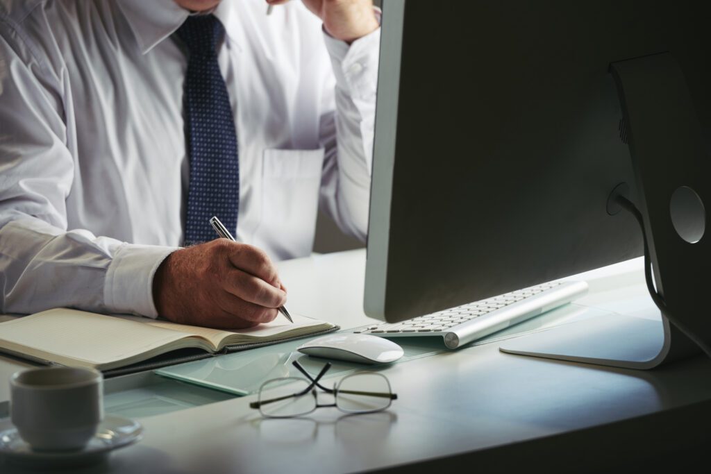 Man writing in notebook at desk with computer