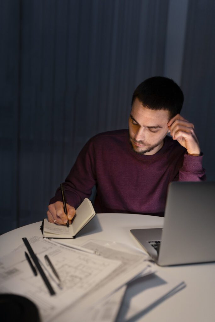 Man writing in notebook at desk with laptop